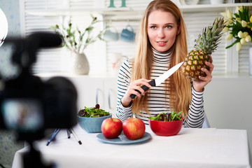 Food blogger cooking fresh vegan salad of fruits in kitchen studio, filming tutorial on camera for video channel. Female influencer holds apple, pineapple and talks about healthy eating. Fructorianism - Powered by Adobe