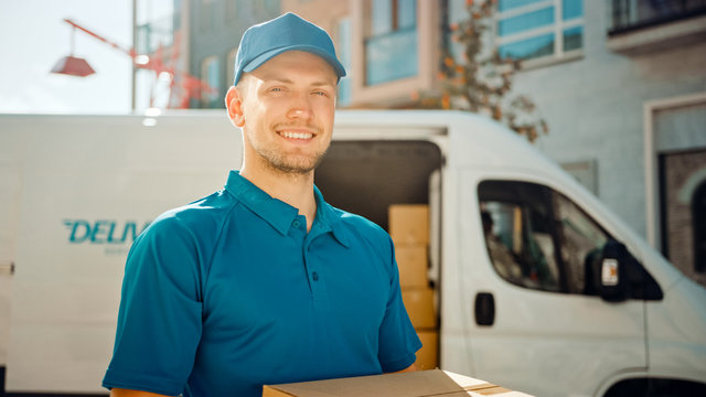 Portrait of Handsome Delivery Man Holds Cardboard Box Package Standing in Modern Stylish Business District with Delivery Van in Background. Smiling Courier On Way to Deliver Postal Parcel to Client