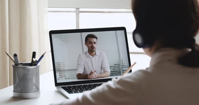 Elearning concept. Over shoulder view of student wearing headphones watching webinar with male trainer, distance learning online looking at laptop screen, writing notes. Remote education training