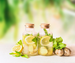 Healthy summer lemonade drinks in bottles on white table at green sunny nature background. Ginger, lemon, mint leaves lemonade. Infused water