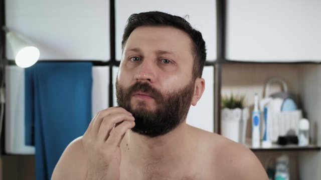 Man is combing his beard. Attractive man in bathroom looks in mirror and combes black hair on his face. Hygiene, hair care, freshness concept. Close-up