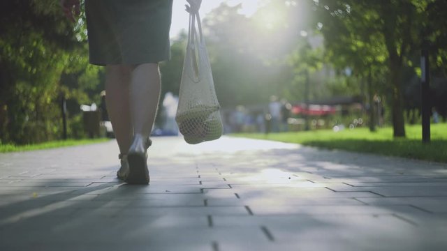 Young woman holds cotton mesh shopping bag with vegetables and walks in the summer park