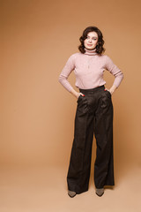 Studio shot of a gorgeous trendy brunette with wavy hair. Model wearing smart clothes - pale pink sweater and dark brown trousers. Posing with hands in pockets.