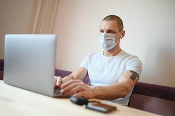 Fototapeta na wymiar Young european man in face protective medicine mask working on a laptop during coronavirus isolation home quarantine. Covid-19 pandemic Corona virus. Distance online work from home concept.