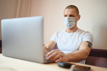 Young european man in face protective medicine mask working on a laptop during coronavirus isolation home quarantine. Covid-19 pandemic Corona virus. Distance online work from home concept.