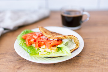 Oat pancake on a white plate stuffed with cream cheese, cheese and tomatoes with green leaf staying on a wooden table with cup of coffee on a background. Healthy food