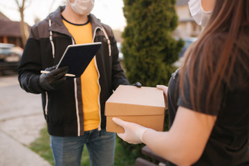 Man from delivery service in medical mask and gloves handing fresh food to young woman customer receiving express delivery from courier at home. Courier with tablet, customer sign in. Female order