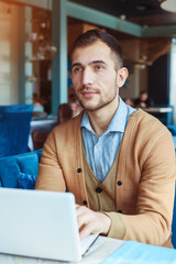 Fototapeta premium A young man works behind a laptop in a cozy cafe. Business and coworking concept. Portrait of a man.