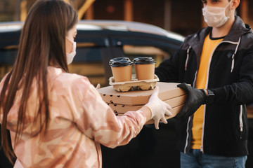 Young woman in protective mask and medical mask take order from delivery boy outside. Food delivery in quarantine. Courier bring Pizza and coffee for customer