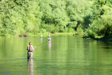 Fly fishing on the river in a rural place in the summer, young fishing woman standing in the water.Unrecognizable people in the background