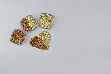 Green and fried dry buckwheat in cans and scattered in the shape of a heart on a gray background, top view with copy space