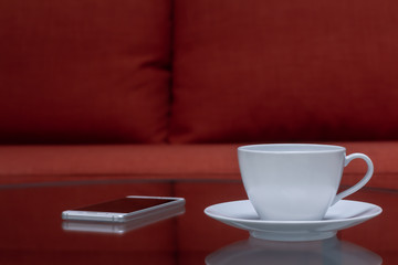 Empty white coffee cup or teacup and coaster for coffee put on clear table near blurred smartphone and red cloth sofa background.