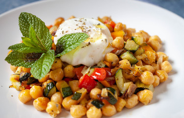 Homemade spicy vegetarian stir fry with chickpeas and pepper and zucchine dice with a fried egg and some mint decoration in a white plate. Close up.