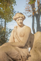 Ancient stone statue of an beautiful angel with wings against blue sky and trees.