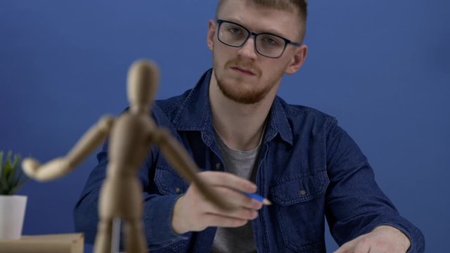 Designer depicts a wooden gestalt figure in creative workspace in blue tones. The work of the artist or designer. The creative process. A man with glasses is copying on paper a wooden man.