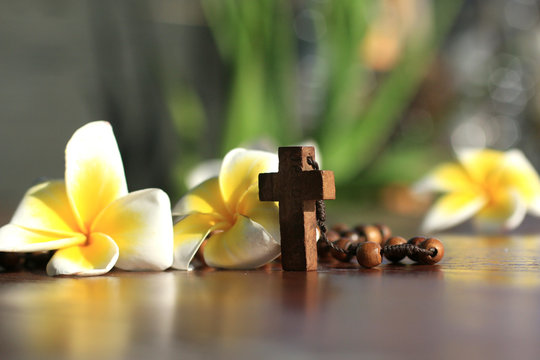 Holy Cross of rosary closeup in low angle view. Wooden rosary with Jesus Christ crucifix  and Bali frangipani flowers. Copy space on the background  for your text design. Catholic spiritual symbol.