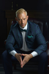 a businessman in a blue jacket sits in a chair, concentrating in a pose like Trump's. on dark...