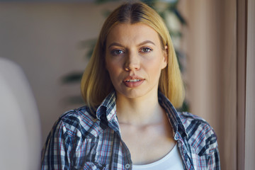 Front view portrait of young beautiful caucasian blonde woman female girl standing by the window wearing shirt at home in day looking to the camera