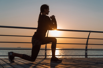 Young athletic girl performs squats on wooden embankment by the sea in early morning. Healthy lifestyle, coaching, training concept.