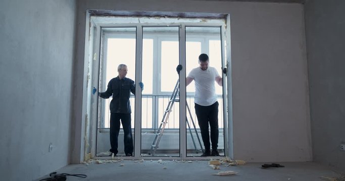 Two men at real estate object under construction dismantle plastic window, cut foam with knives, repair new apartment, replace window block, dusty work, builders, opposite window, construction site