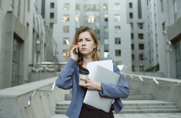 Fototapeta premium Puzzled businesswoman in front of office building having phone conversation holding laptop and documents.Female office worker with puzzled expression dealing with deadlines holding papers and computer