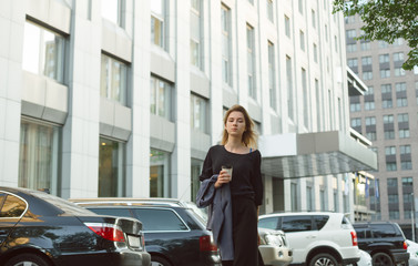 Naklejka na ściany i meble Portrait of stylish young girl dressed in smart casual clothes holding jacket strolling on street of modern city.Young woman walking in urban setting looking at camera near the office and car parking.