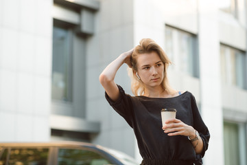 Thoughtful woman listening to music with wireless headphones holding coffee in the urban scenery. Pensive young girl looking aside in her earphones holding take away coffee cup. Office worker concept.