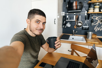 Handsome man taking selfie while working on laptop at home in modern loft kitchen. Remote work.