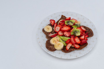 Brown homemade pancakes with fresh fruits, strawberries, bananas, kiwi and with honey on a light plate. Healthy food, gluten free, no eggs. Copy space. White background. Flat lay. Top view.