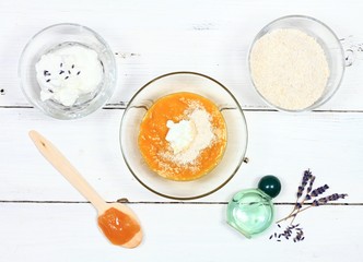 Homemade face mask from mango, heavy cream, finely ground oats and honey. Three bowls full of food ingredients and lavender oil on the white table, flat lay