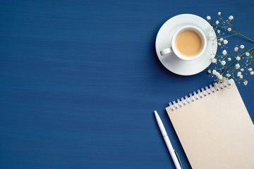 Feminine workspace with cup of coffee, paper notepad and flowers on classic blue background. Flat...