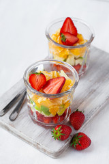 Concept of healthy food, clean eating. Low calories sweet summer dessert. Homemade simple fruit salad in two glasses for breakfast. Orange, apple, strawberry. White wooden background, copy space