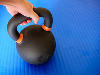 Hand holding black kettlebell on blue gym mat. Home workout concept