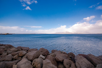 Scenic landscape shot of calm ocean waters shot from a shore with big rocks in foreground. The blue skies & white clouds are visible till horizon with an island at far end. Peaceful & Tranquil shot.