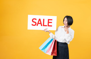 Young smiling asian woman holding multi coloured shopping bags and hands showing sale sign on light orange background.