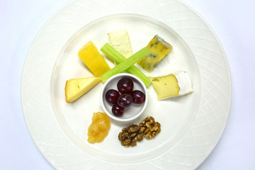 A selection of cheeses with grapes, chutney, celery & walnuts all prepared to serve as a cheese plate, on a white table cloth.
