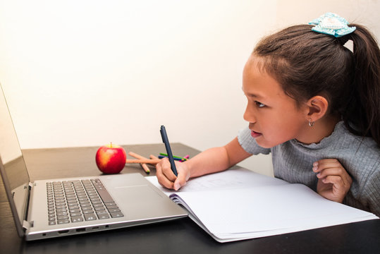 Little girl writing on a notebook. Doing homework at home with the laptop. Pretty little girl studying and learning at home during the quarantine of the coronavirus infection.