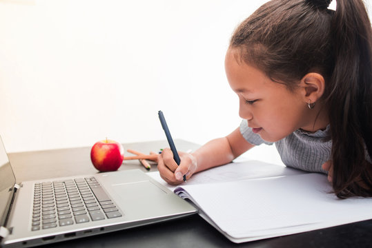 Little girl writing on a notebook, taking class from the laptop on white background. Concept of online classes at home during the quarantine.