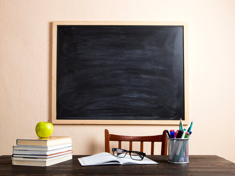 Books, apple, pens, pencils and glasses on a wooden table, against the background of a chalk board. Concept for Teacher's Day, copy space.