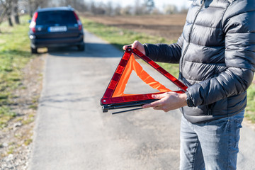 young man installs an orange warning triangle on the road in front of a broken car.