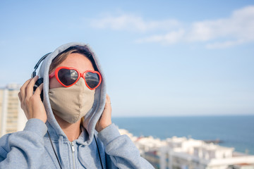 Female wearing protective mask and red love heart shape sunglasses using headphones listening music on outdoors background. Message of hope, having fun creative self-isolation. Closeup portrait person