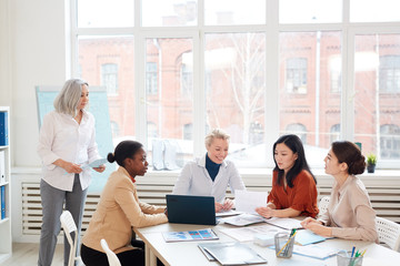 Wide angle view at diverse group of businesswomen discussing project while sitting at table against window during meeting in conference room, copy space
