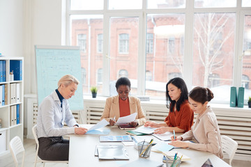 Diverse group of successful businesswomen discussing project while sitting at table against window during meeting in conference room, copy space