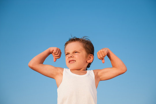 Strong Little Caucasian Boy In White Tank Top Showing Strong Muscle On Blue Sky Background