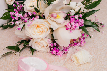 Wedding bouquet of cream and pink roses, ruskus leaves, pink gypsophila and decorative ribbons for decoration of the bouquet are on a white table.
