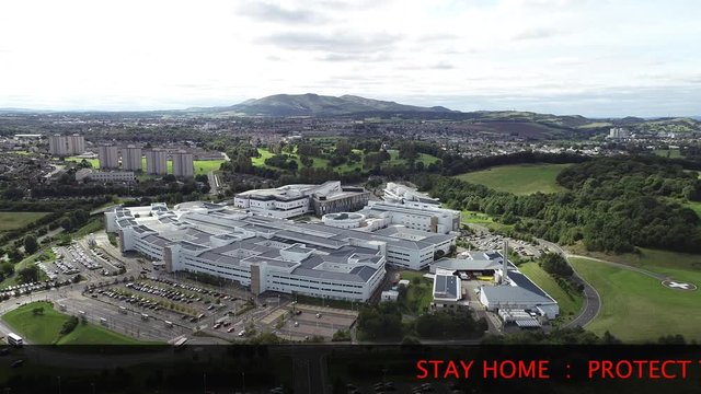 Aerial footage of the Edinburgh Royal Infirmary and the new Royal Hospital for Sick Children at Little France. Flying away, with text scrolling at the bottom; STAY HOME : PROTECT THE NHS : SAVE LIVES.