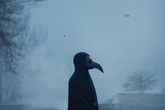 Dark black figure of a man in a mask of plague doctor stands in the dusk on fog cemetery