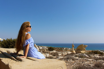 a young beautiful girl with glasses in a blue dress with long unpainted hair sits on a stone slab and looks at the blue surface of the sea and the clear sky. Recreation and tourism in Cyprus.