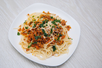 Spaghetti with minced meat in tomato with herbs and cheese on a white plate. Dish with food on a light wooden table. Lunch with meat.