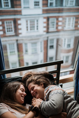 Girls couple with long light brown hair lying on a bed in front of a window overlooking the city.